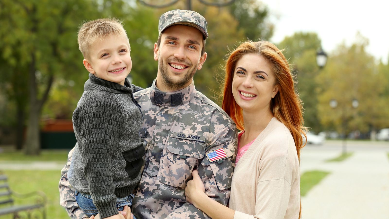 Service member holding his young son next to his smiling wife on a tree-lined street
