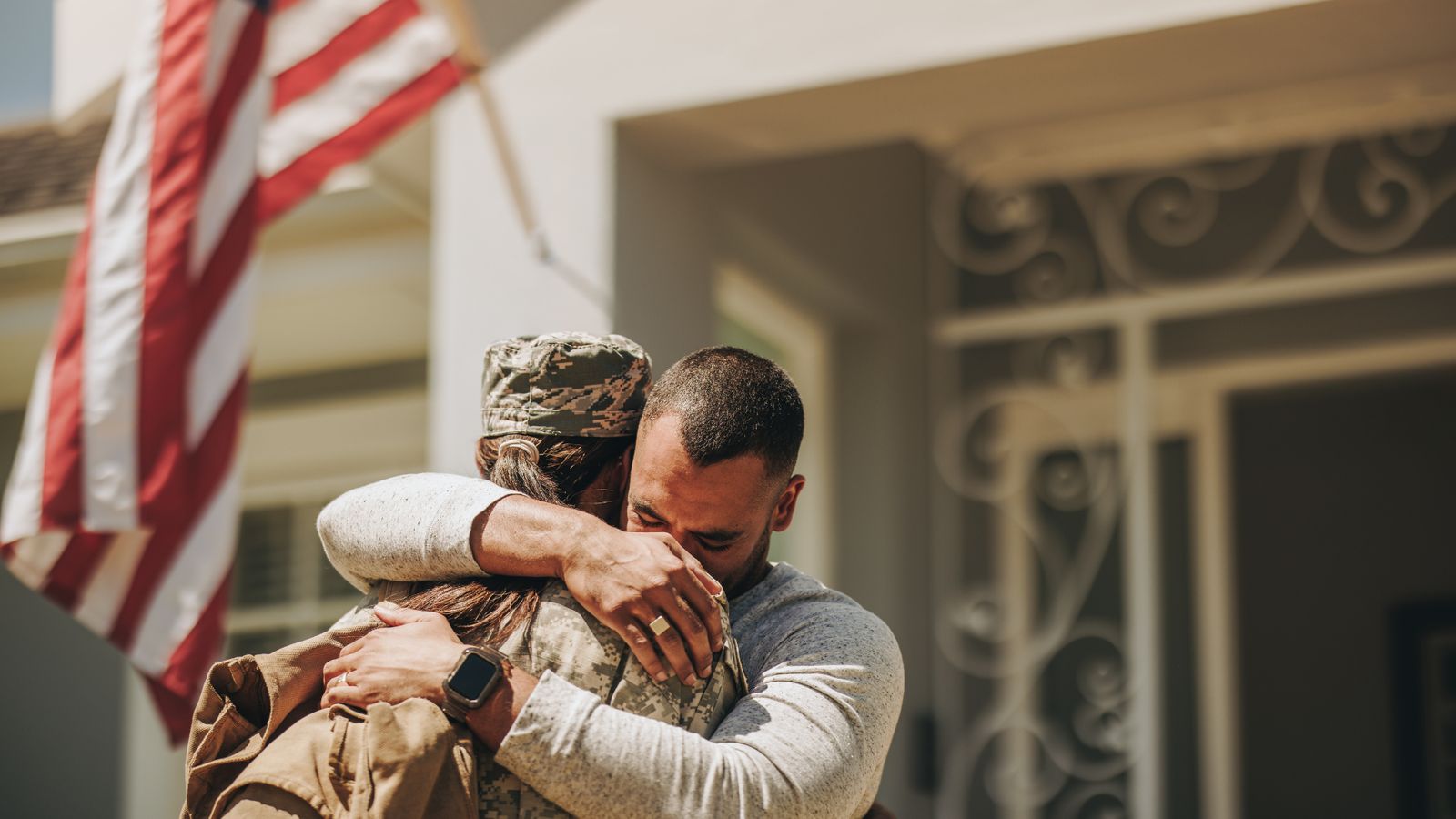 Service member embracing a loved one on a front porch beside an American flag