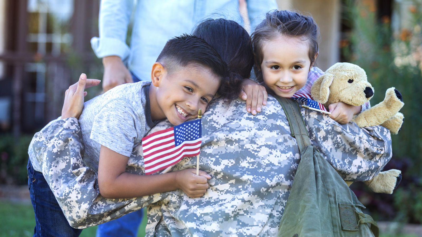 Two children hugging a service member, one waving an American flag