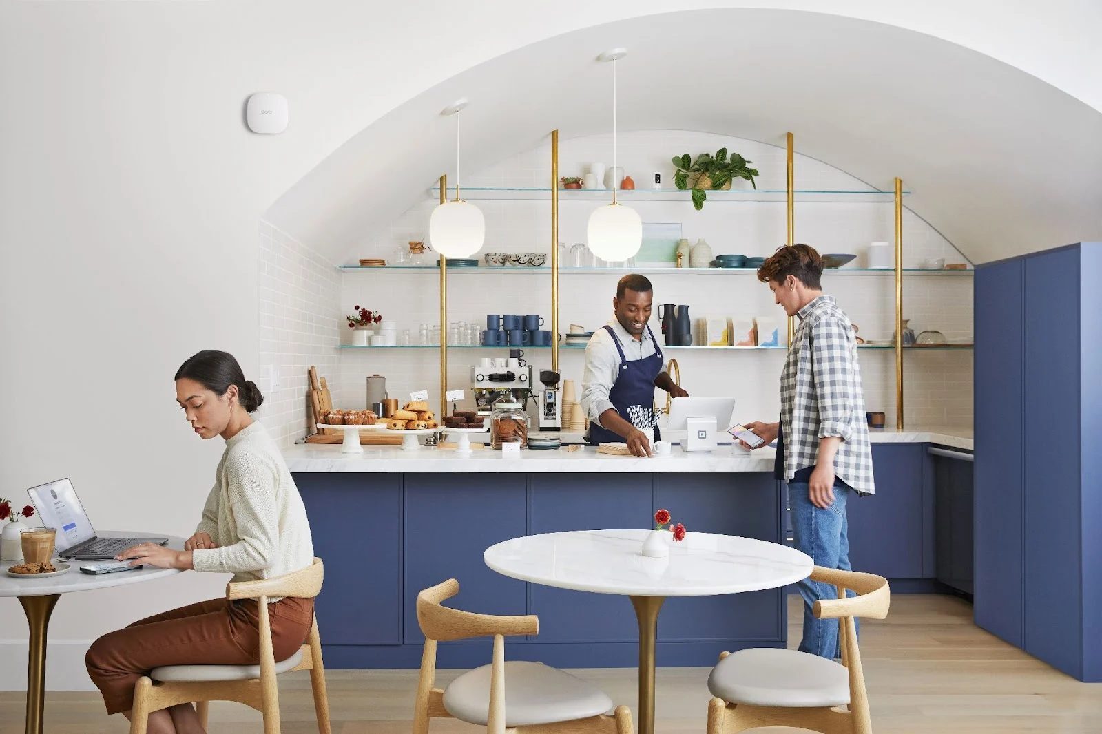 A barista helps a customer at a cafe counter while a guest works on a laptop, an eero access point mounted on the wall