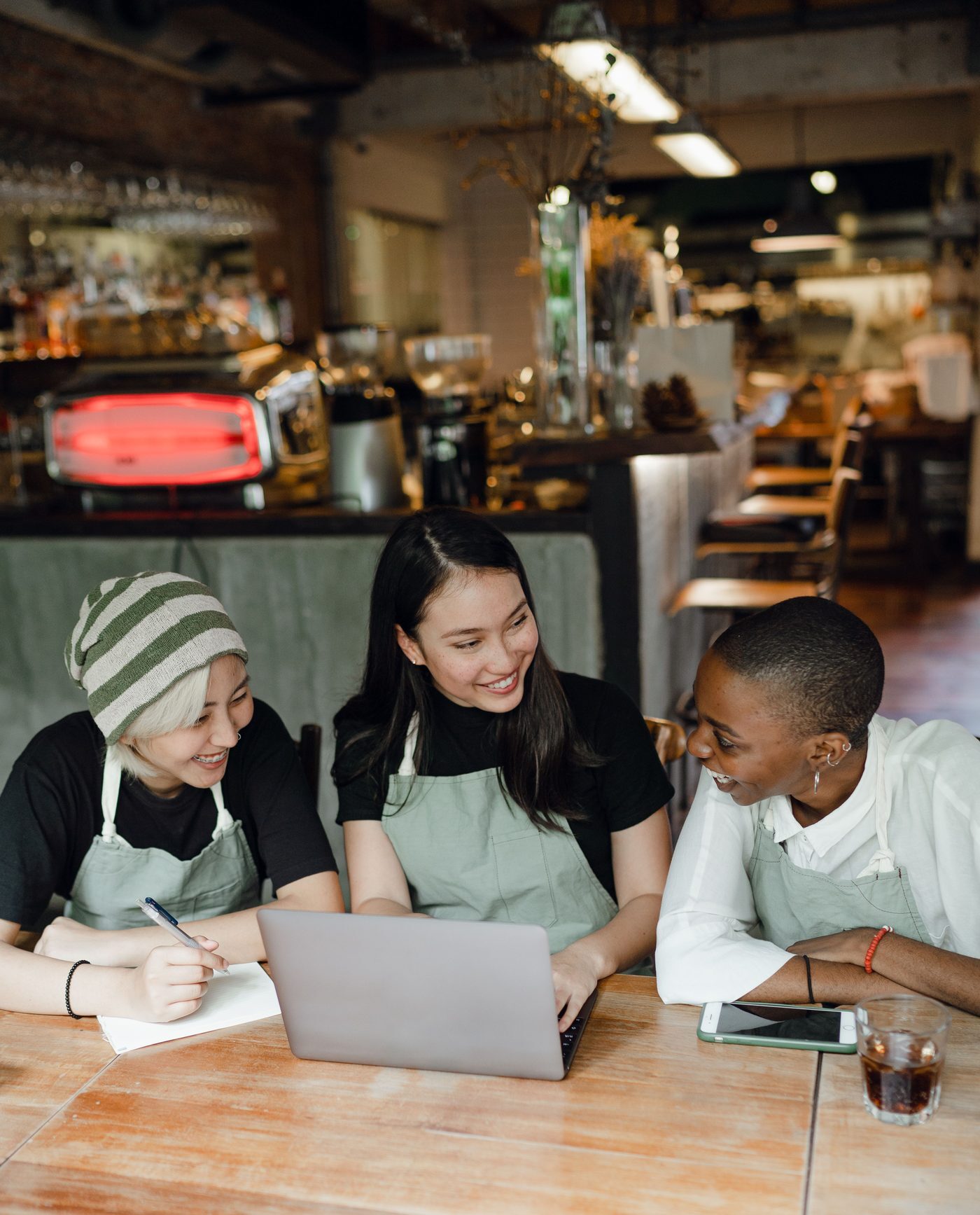 Three cafe team members in aprons smile while checking a laptop at a wooden table