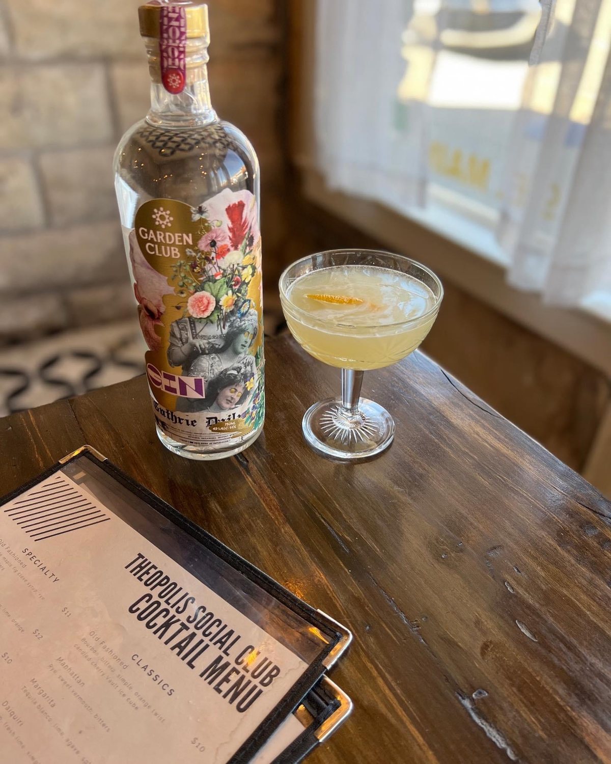 A gimlet in a coupe beside a bottle of Garden Club Gin and the Theopolis Social Club cocktail menu